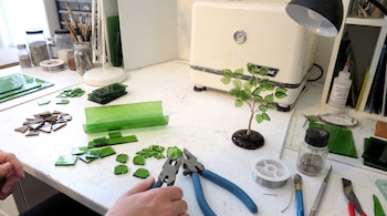 Manos con alicates junto a fragmentos de vidrio verde, alambre, herramientas de corte y un horno sobre una mesa de trabajo.