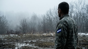Hombre de espaldas con chaqueta de camuflaje y parche de la bandera de El Salvador frente a un campo con lodo y árboles entre la niebla.