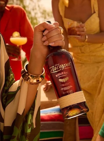 Mano de persona con uñas pintadas sostiene una botella de Ron Zacapa. Brazalete dorado en la muñeca. Al fondo, personas con bebidas y un tejido de rayas