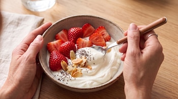 Dos manos sostienen un tazón cerámico con yogur, fresas en rebanadas, almendras y semillas de chía sobre una mesa de madera.