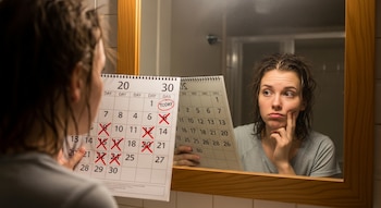 Mujer de espaldas, frente a un espejo, sostiene un calendario con días marcados con cruces y un círculo rojo. Su reflejo muestra cabello graso.