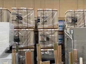 Vista de un almacén industrial con pilas de cajas y electrodomésticos empaquetados en plástico transparente sobre palets; listones de madera en primer plano