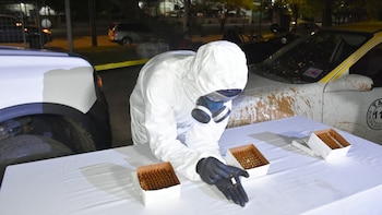 Uma pessoa com traje de proteção individual branco, máscara de gás e luvas pretas olha para um frasco âmbar. Três caixas cheias de pratos sobre uma mesa branca e dois carros sujos ao fundo