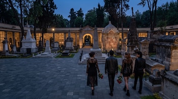 Cuatro personas vestidas de negro caminan de espaldas sosteniendo rosas rojas entre lápidas y monumentos en un panteón histórico al atardecer.
