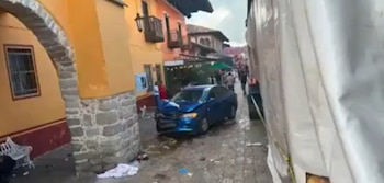 Coche azul con frontal dañado en calle estrecha y mojada. A la izquierda, arco de piedra y edificios. A la derecha, un camión blanco. Personas al fondo