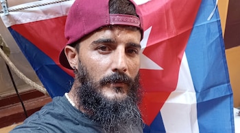Un hombre con barba, gorra roja y un collar blanco se encuentra ante una bandera de Cuba.