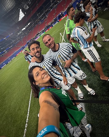 Selfi de mujer sonriente con dos futbolistas en camiseta de rayas celestes y blancas, uno sostiene un trofeo. Otros jugadores y estadio en Atlanta