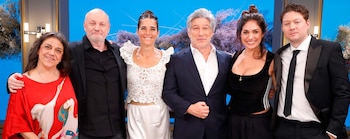 Seis personas, tres hombres y tres mujeres, posan sonriendo frente a un fondo azul con decoraciones florales. Todos visten ropa formal o semiformal