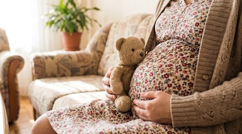 Primer plano de una mujer embarazada sentada, vestida con un vestido floral y cárdigan. Sostiene un osito de peluche contra su vientre, con una mano sobre este.