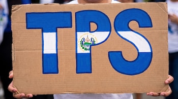 Manos sostienen un cartel de cartón con las siglas TPS pintadas en azul y blanco. La letra P contiene el escudo nacional de El Salvador.