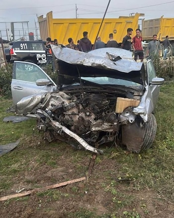 Coche plateado severamente dañado con el capó abierto en campo de hierba. Se observan personas, un camión volquete amarillo y un vehículo policial 911