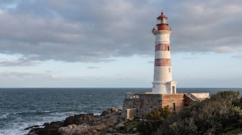 Faro blanco y rojo con franjas sobre un terreno rocoso, con el mar azul oscuro y un cielo nublado al fondo.