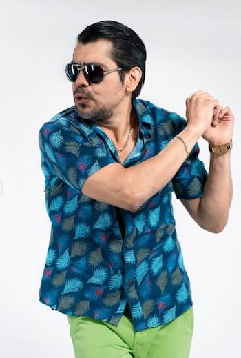 Pietro Sibille con gafas de sol, camisa azul de hojas tropicales, pantalones verde lima, barba y pelo oscuro, posando con las manos levantadas
