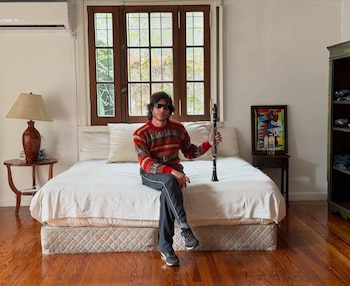 Un hombre con gafas oscuras y suéter estampado está sentado sobre una cama con un clarinete en la mano frente a una ventana