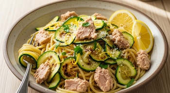 Un plato de espaguetis con rodajas de calabacín, trozos de atún, queso rallado, perejil, ralladura de limón y dos gajos de limón. Un tenedor metálico en la pasta.