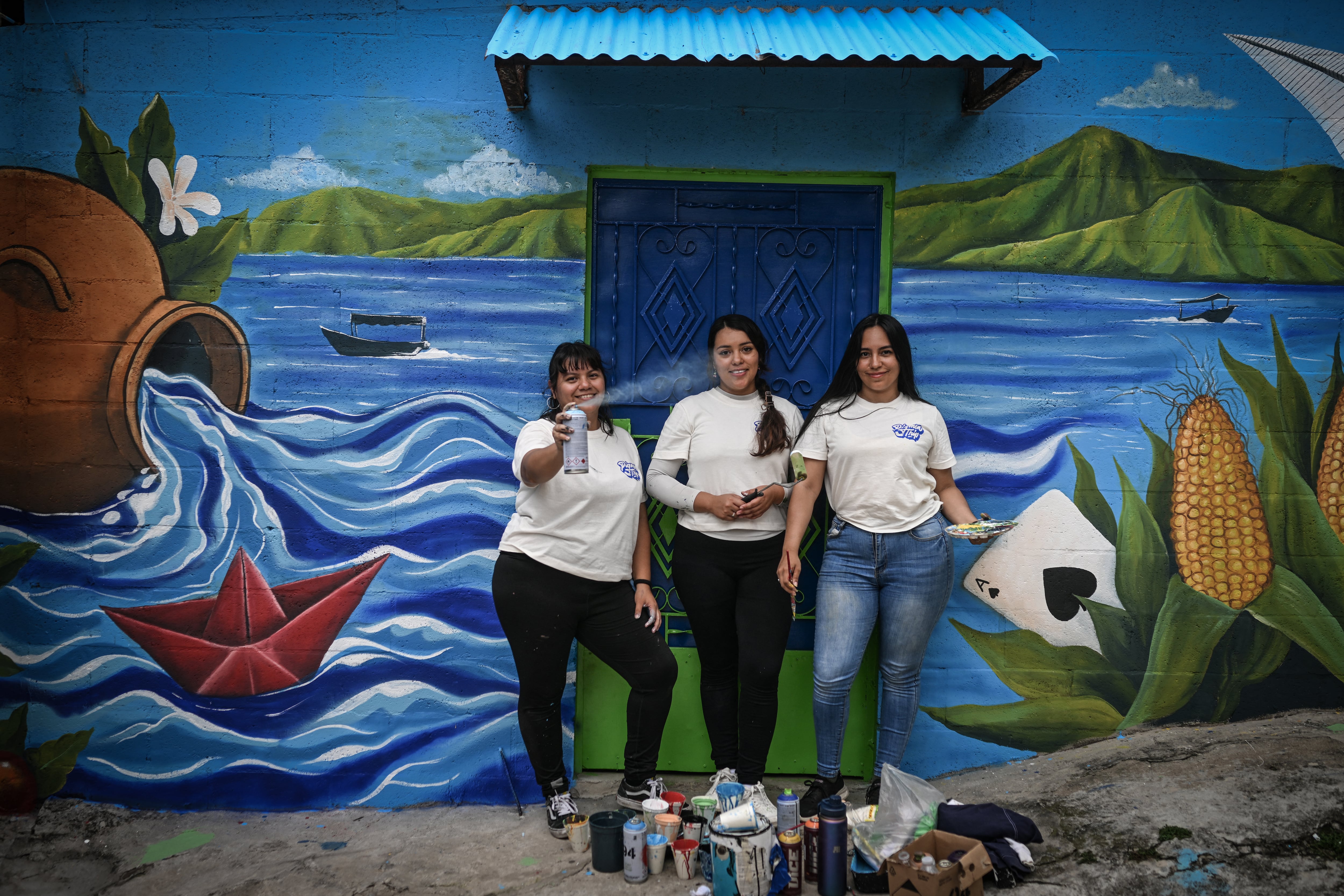 Las artistas salvadoreñas Patricia Vega (izq.), Elizabeth Guardado (centro) y Marcela Alvarenga (der.) posan frente a una casa cuya fachada están decorando con el mural