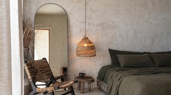 Dormitorio con cama de sábanas verde oliva, espejo de cuerpo entero arqueado, silla tejida, lámpara colgante de ratán y pared texturizada.