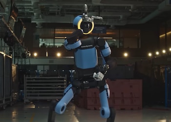Robot humanoide Boston Dynamics azul y negro con un anillo de luz amarilla en la cabeza, realizando un movimiento en un almacén