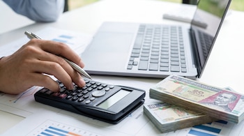 Primer plano de una mano utilizando una calculadora en una mesa, con dos pilas de billetes de Quetzales y un ordenador portátil al fondo.