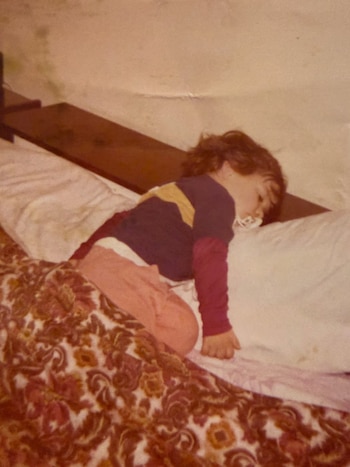 Un niño pequeño duerme de lado en una cama, con un chupete en la boca, vistiendo una camiseta y un pantalón, cubierto por una manta estampada
