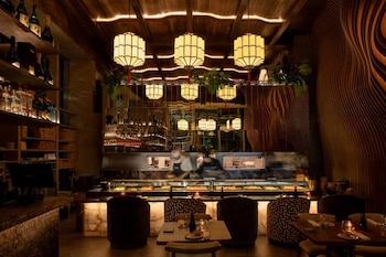 Interior de un restaurante con barra iluminada, cocineros detrás del mostrador, mesas, sillas y lámparas colgantes de estilo asiático