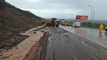 Una pala mecánica amarilla limpia barro en una carretera mojada junto a vehículos y una persona con impermeable