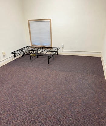 Habitación vacía con paredes claras, alfombra oscura, ventana con persianas y una estructura metálica para cama en la esquina