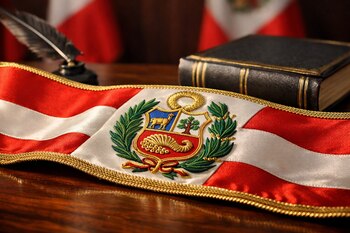 Banda presidencial de Perú roja y blanca con bordado del escudo nacional, un libro oscuro, una pluma y una mesa de madera. Banderas peruanas al fondo.