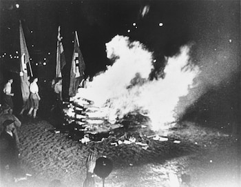 Fotografía en blanco y negro de una gran hoguera nocturna con llamas intensas y humo. Libros arden en el fuego, y varias banderas con esvásticas son visibles