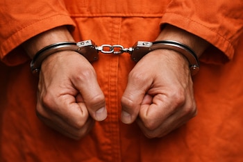 Primer plano de las manos de una persona con esposas de metal, vestida con un uniforme penitenciario naranja, mostrando las muñecas y parte del brazo.
