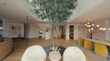 Gran salón interior con árbol, escaleras, sillones blancos, sofá curvo, mesa de comedor con sillas amarillas y lámpara de techo