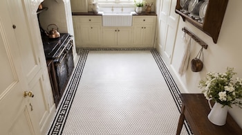 Cocina con piso de mosaico hexagonal blanco y negro, gabinetes blancos, fregadero de granja, estufa con tetera de cobre, estante con platos, perchero con toalla, mesa con flores.