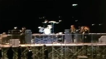 Integrantes de una banda musical tocan instrumentos sobre un escenario elevado iluminado de noche y protegido por vallas