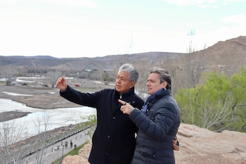 Dos hombres con abrigo oscuro de pie sobre rocas señalan hacia un río y cerros en el fondo