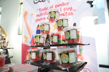 Vasos transparentes de gomitas rojas y verdes de la marca GOMICUY, exhibidas en un soporte negro de varios niveles frente a un cartel borroso