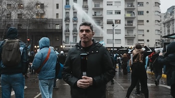Un hombre con chaqueta oscura sostiene un micrófono en una calle mojada con edificios, personas y un chorro de agua al fondo