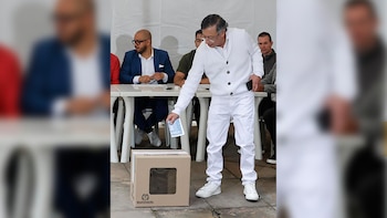 Un hombre con camisa blanca y pantalón blanco, lentes y cabello gris, deposita una papeleta en una urna de cartón con una ventana