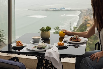 Una persona sentada en una mesa de desayuno con vista al mar, sosteniendo un vaso de jugo. Sobre la mesa hay platos con waffles, huevos pochados, pasteles y café