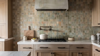 Una cocina con gabinetes de madera clara, encimeras de mármol blanco, un backsplash de azulejos cuadrados verdes y beige, una estufa y una campana extractora.