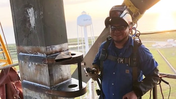 Un soldador sonriente con casco, gafas de sol y arnés trabaja en una estructura metálica. Al fondo, una torre de agua "SPAC" y paisaje de Starbase al atardecer