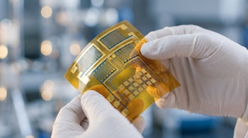 Primer plano de manos con guantes de látex blancos sosteniendo un sustrato flexible dorado con patrones de circuitos electrónicos azules y dorados.