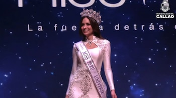 Una mujer de cabello castaño oscuro con una corona brillante y un vestido blanco adornado con perlas, luciendo la banda de Miss Perú Universe, sonríe en un escenario oscuro