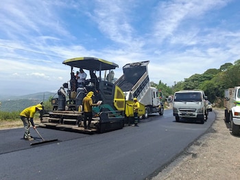 Una máquina pavimentadora, un camión volcador descargando asfalto, varios obreros con chalecos amarillos y otros vehículos en una carretera