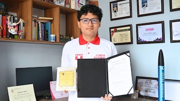 Joven de cabello oscuro y gafas, con polo blanco con la bandera de Perú, sonríe sosteniendo dos diplomas frente a una estantería y certificados enmarcados