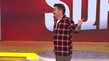 Migue Granados en un escenario, vestido con camisa a cuadros roja y negra, sobre un fondo rojo y piso amarillo, gesticulando con la mano derecha