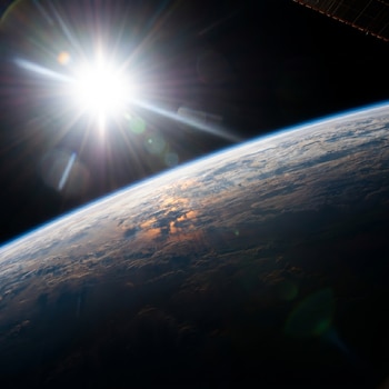 Vista de la Tierra desde el espacio mostrando el horizonte curvo, nubes y un sol brillante con destellos. Un panel solar es visible en la esquina superior derecha.