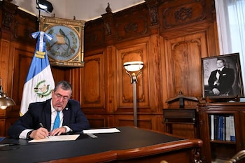 Un hombre con traje oscuro y corbata azul firma un papel sentado ante un escritorio de madera, al lado de una bandera de Guatemala