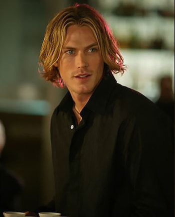 Jason Lewis, hombre de cabello rubio ondulado hasta los hombros y ojos azules, viste una camisa negra abotonada y mira ligeramente hacia la derecha