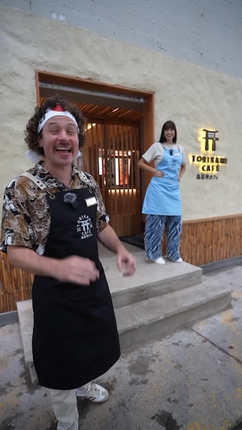 Luisito Comunica sonríe y gesticula con una camisa estampada y delantal negro; su pareja sonriente en un delantal azul en la entrada del Torikami Cafe