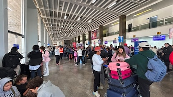 Interior de terminal de aeropuerto con multitud de pasajeros, maletas, mostradores de aerolíneas, pantallas de información y grandes ventanales